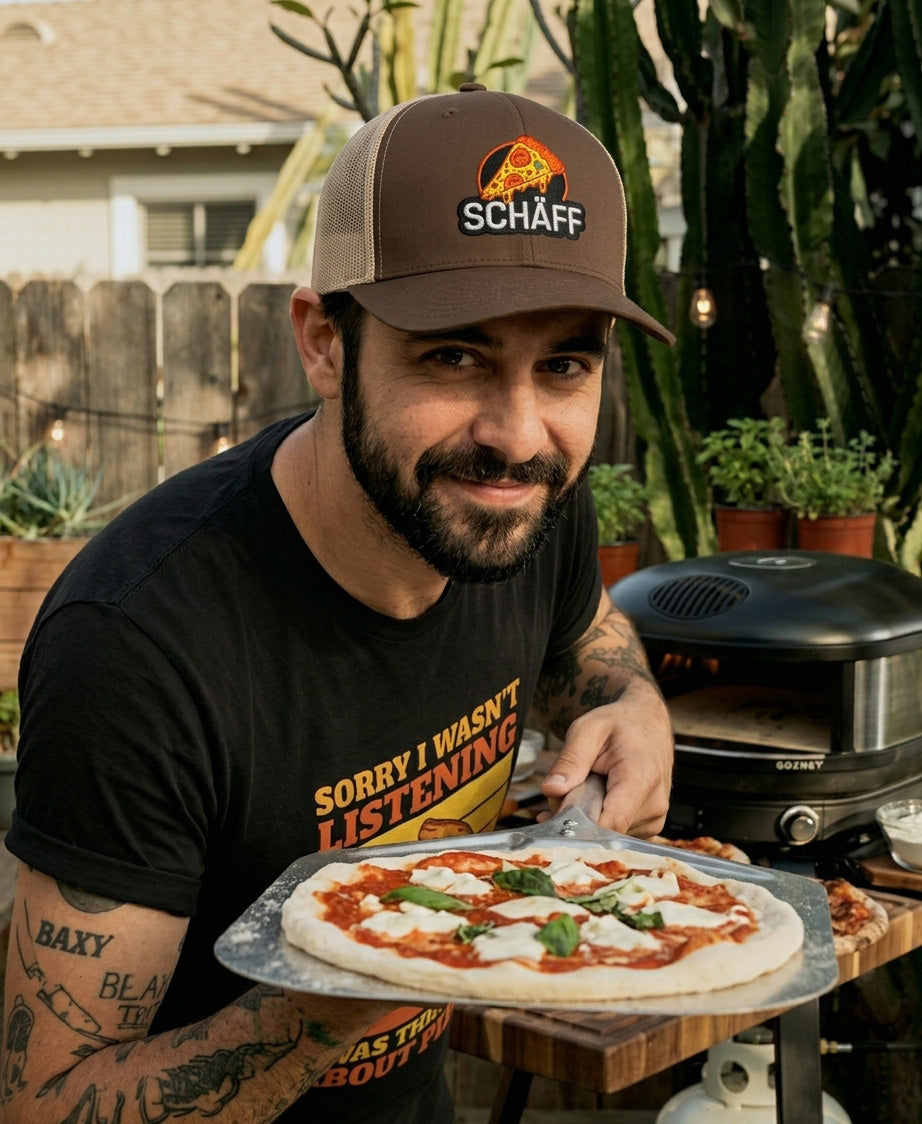 Mann mit Schäff Pizza Retro Trucker Cap hält eine frischgebackene Pizza in die Kamera, im sommerlichen Garten mit Pizzaofen und Kräutern.