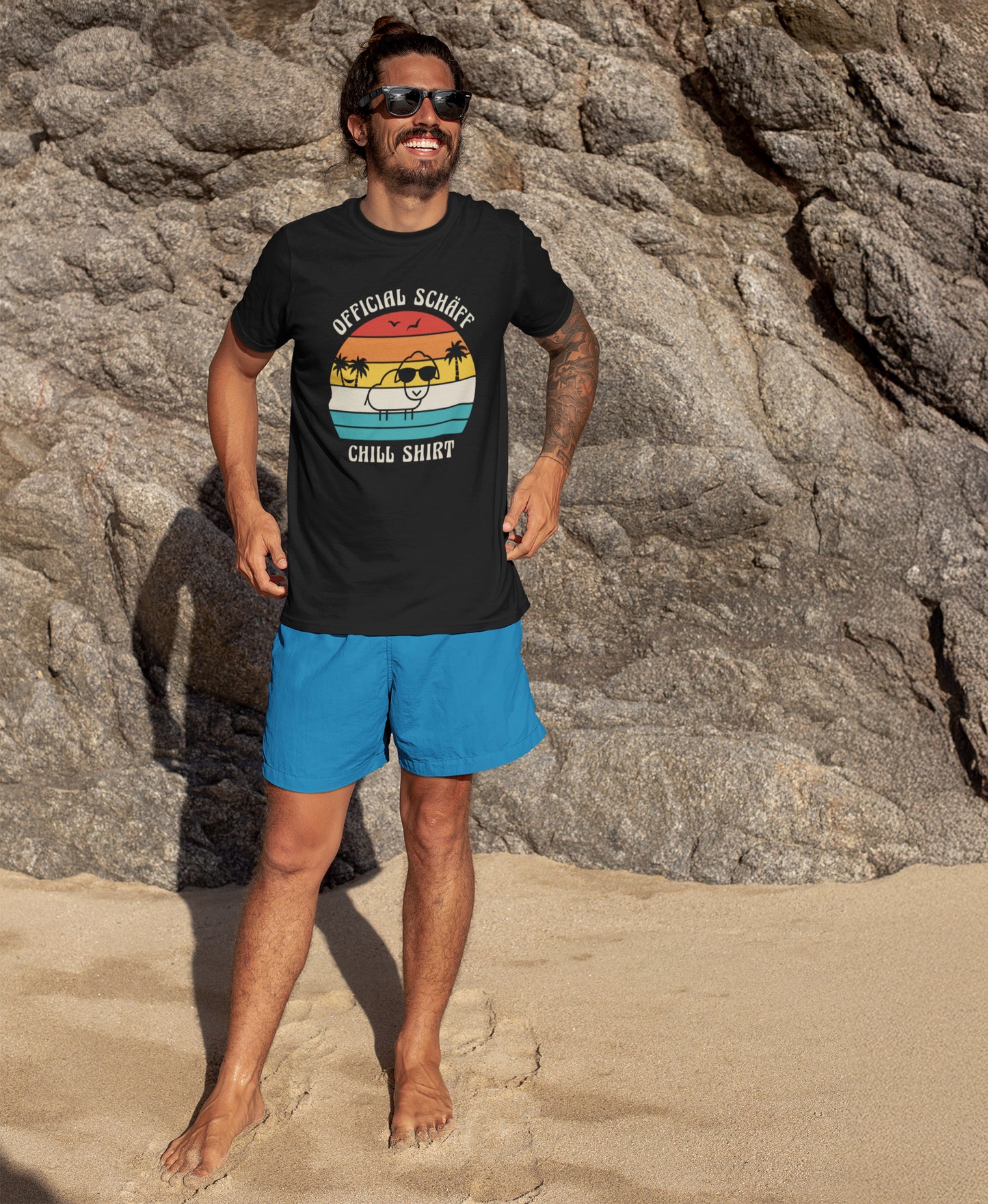 Mann am Strand trägt SCHÄFF – Official Chill Shirt  Bio T-Shirt in Schwarz, mit lustigem Schaf mit Sonnenbrille, Palmen und Retro Streifen in Sunset Farben