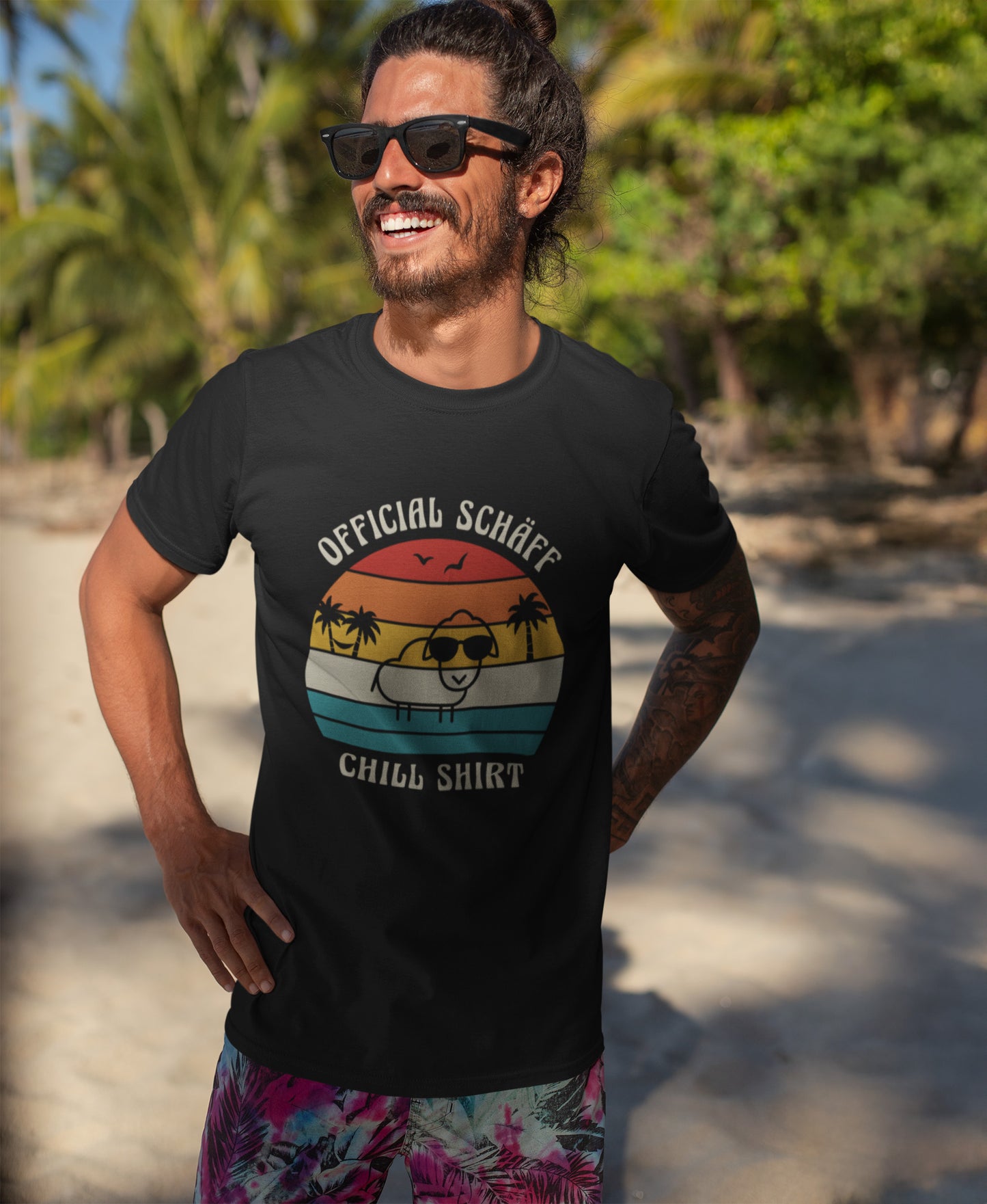 Lachender Mann am Strand trägt SCHÄFF – Official Chill Shirt  Bio T-Shirt in Schwarz, mit lustigem Schaf mit Sonnenbrille, Palmen und Retro Streifen in Sunset Farben