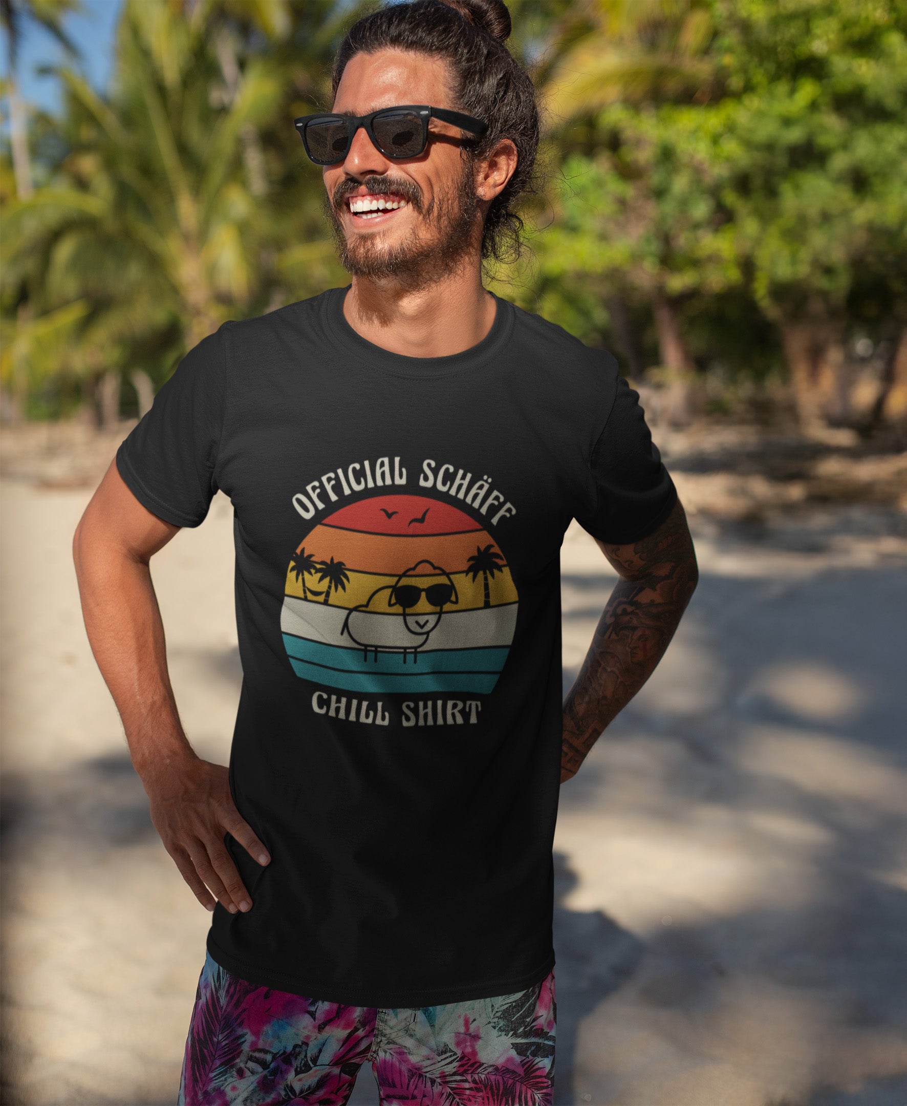Lachender Mann am Strand trägt SCHÄFF – Official Chill Shirt  Bio T-Shirt in Schwarz, mit lustigem Schaf mit Sonnenbrille, Palmen und Retro Streifen in Sunset Farben