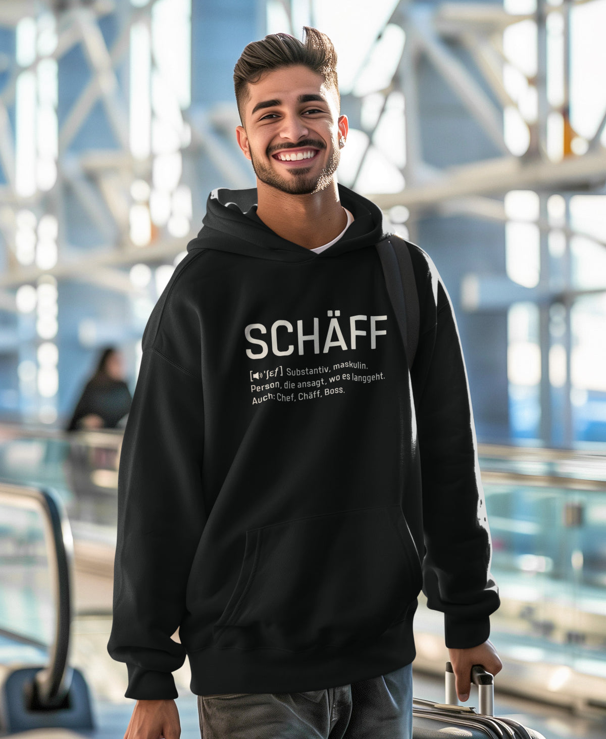 Lachender Mann am Flughafen trägt SCHÄFF – Chef Definition Bio-Hoodie in Schwarz