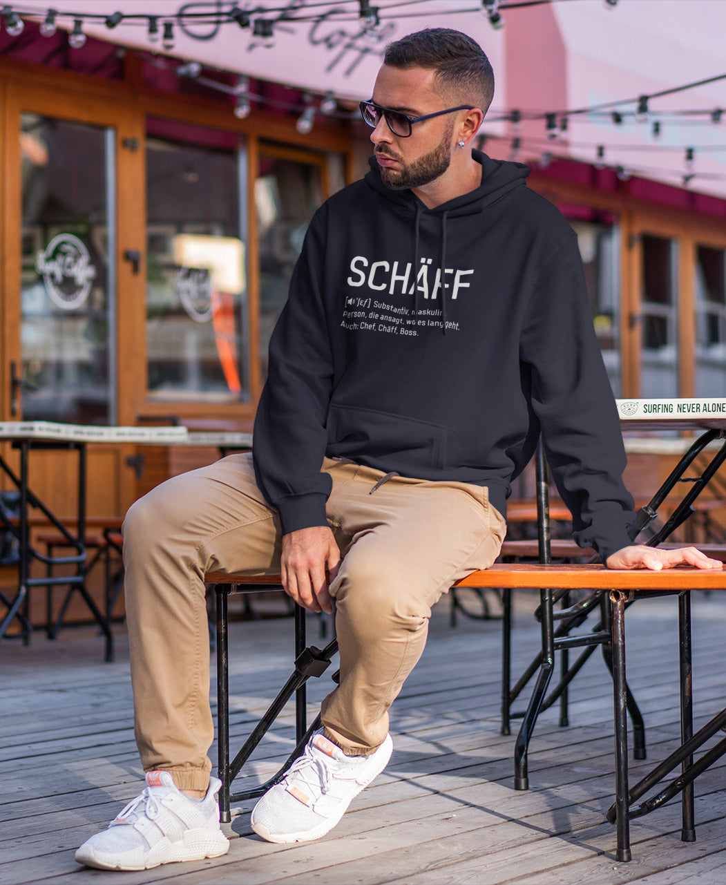Bärtiger Mann mit Sonnenbrille sitzt auf Bank vor Festzelt und trägt SCHÄFF – Chef Definition Bio-Hoodie in Navy