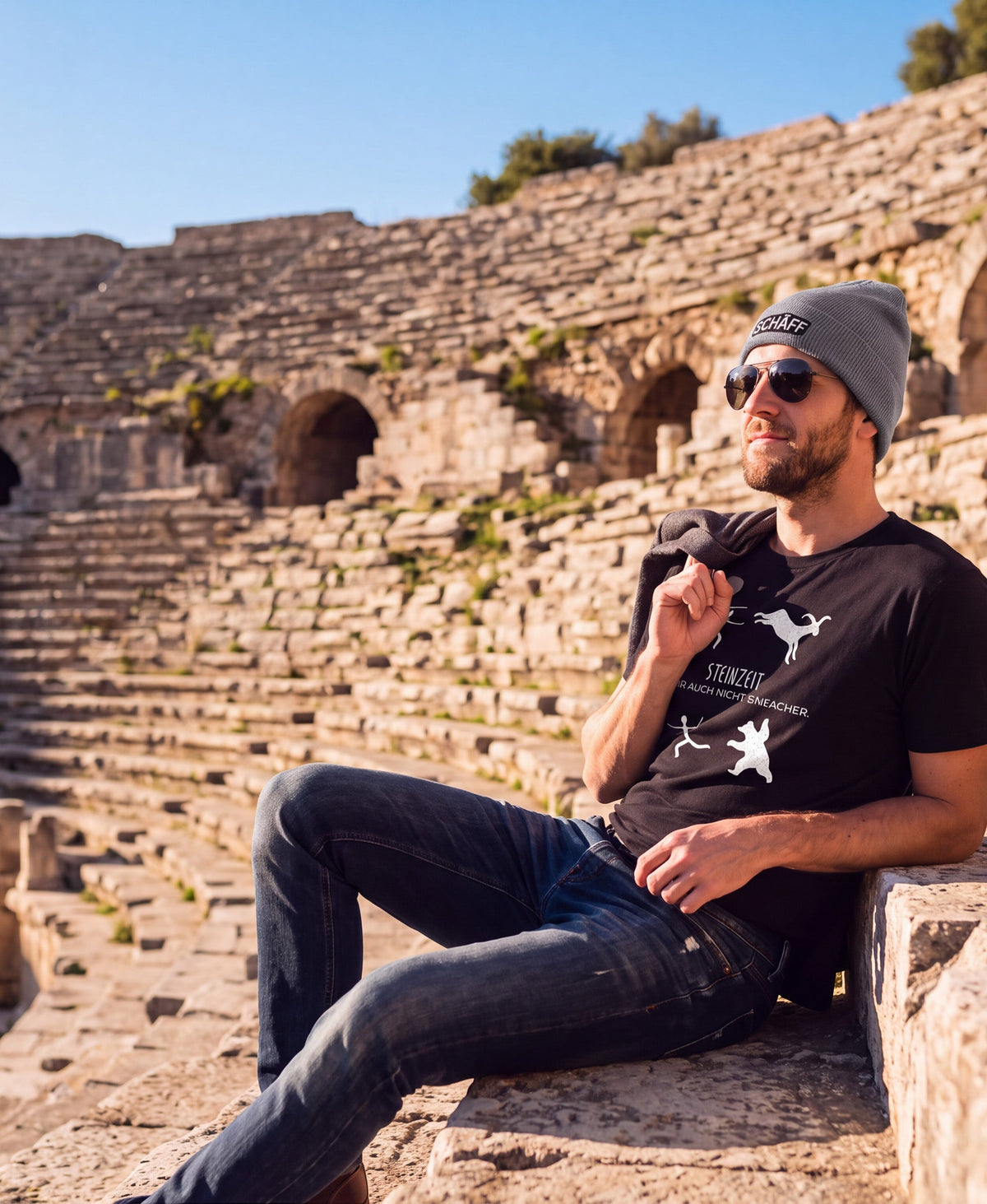 Mann sitzt im Amphitheater mit Sonnenbrille und stylischer SCHÄFF Beanie – Bio-Baumwolle Mütze in Hellgrau mit hochwertiger Stickerei und SCHÄFF – Steinzeit war auch nicht einfacher Bio T-Shirt