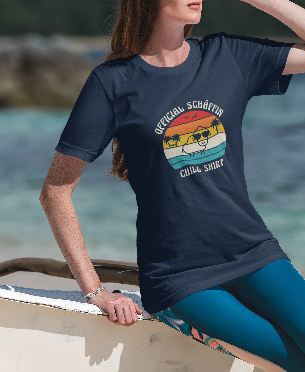 Frau sitzt auf Boot und trägt SCHÄFFIN – Official Chill Shirt  Bio T-Shirt in Navy, mit lustigem Schaf mit Sonnenbrille, Palmen und Retro Streifen in Sunset Farben