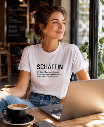 Frau in weißem SCHÄFFIN Chefin Definition Bio T-Shirt mit minimalistischem schwarzem Schriftzug, entspannt mit Laptop in einem Café.