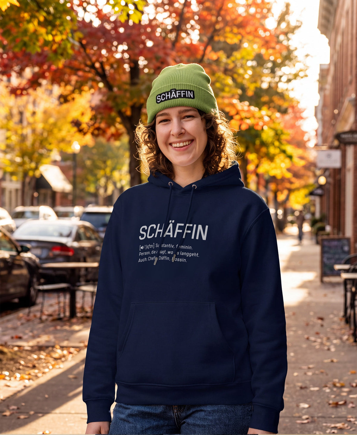 Frau mit SCHÄFFIN Beanie in Grün – Bio-Baumwolle Mütze mit Stick und Frau mit Kaffeetasse und SCHÄFFIN – Chefin Definition Bio-Hoodie in Farbe Navy