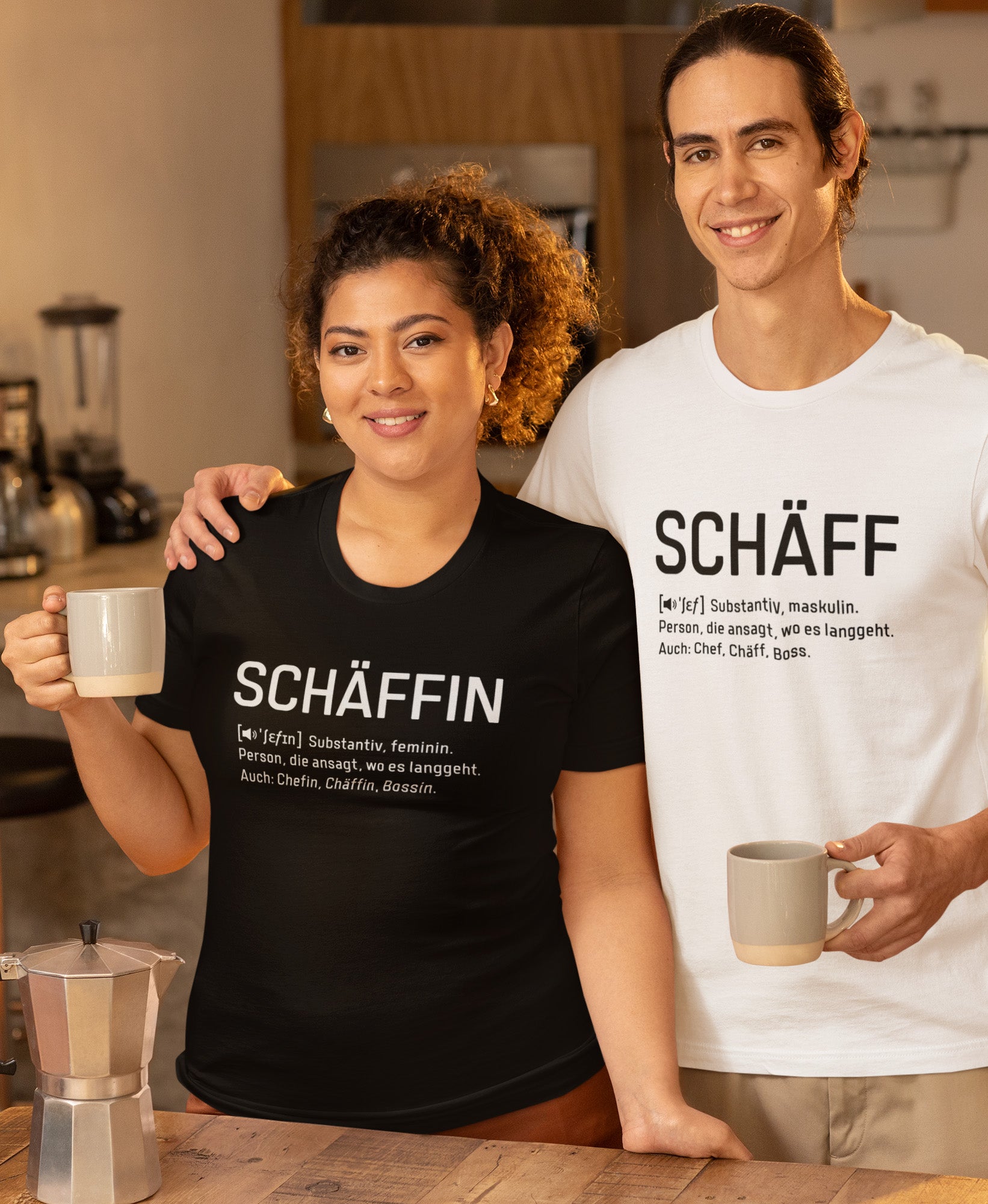 Frau mit Kaffeetasse im Cafe trägt Schwarzes SCHÄFFIN – Chefin Definition Bio T-Shirt mit weisser Aufschrift. Dahinter Mann mit Kaffeetasse trägt weisses SCHÄFF – Chef Definition Bio T-Shirt mit schwarzer Aufschrift.