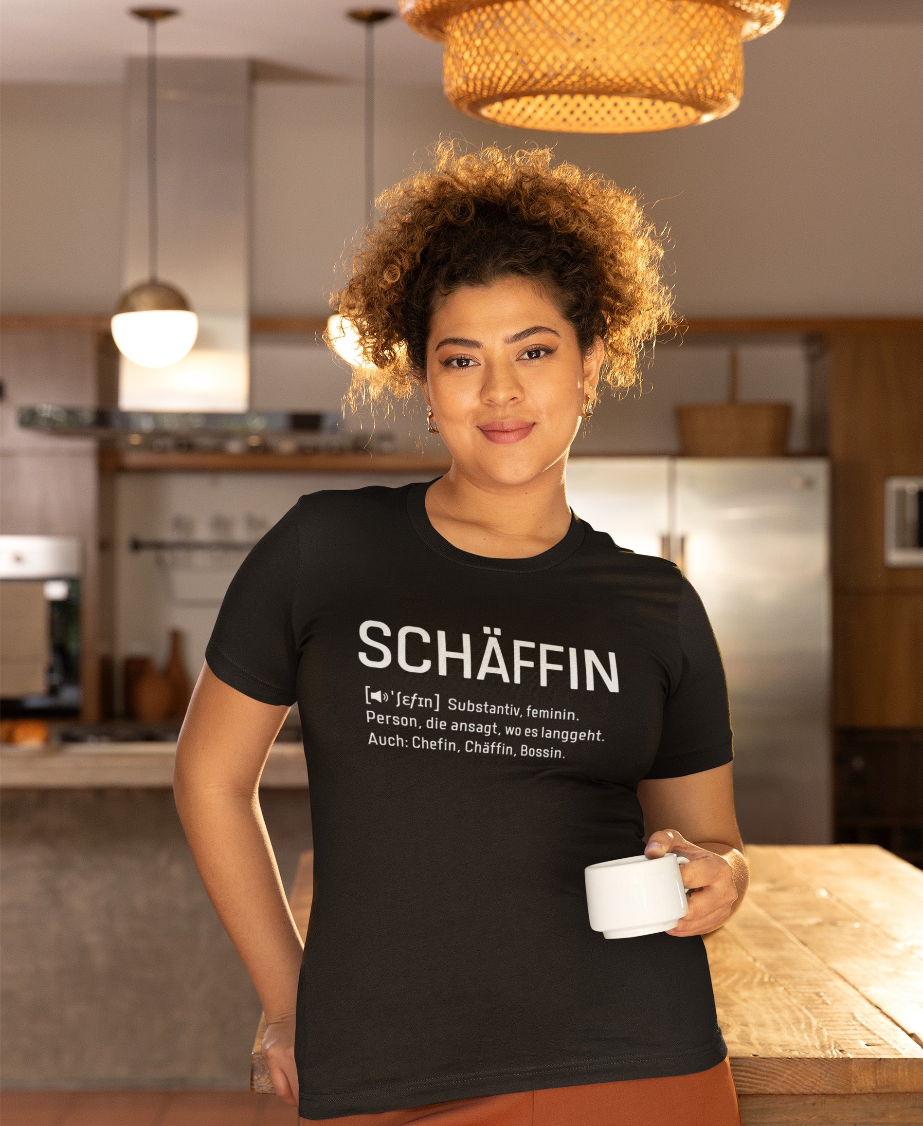 Frau mit Kaffeetasse im Cafe trägt Schwarzes SCHÄFFIN – Chefin Definition Bio T-Shirt  mit weissem Frontaufdruck im Wörterbuch-Stil: SCHÄFFIN – Substantiv, feminin. Person, die ansagt, wo es langgeht. Auch: Chefin, Chäffin, Bossin.