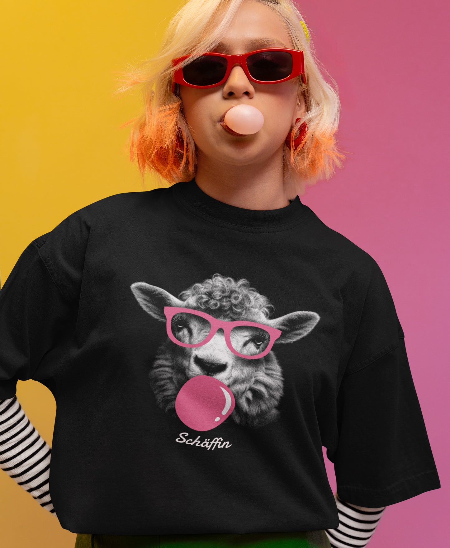 Junge Frau mit trendiger Sonnenbrille und pinkfarbenem Kaugummi, trägt schwarzes SCHÄFFIN T-Shirt mit lustigem Schaf mit rosa Kaugummi und rosa Brille. Darunter ein dezenter Schäffin Schriftzug in Schreibschrift.