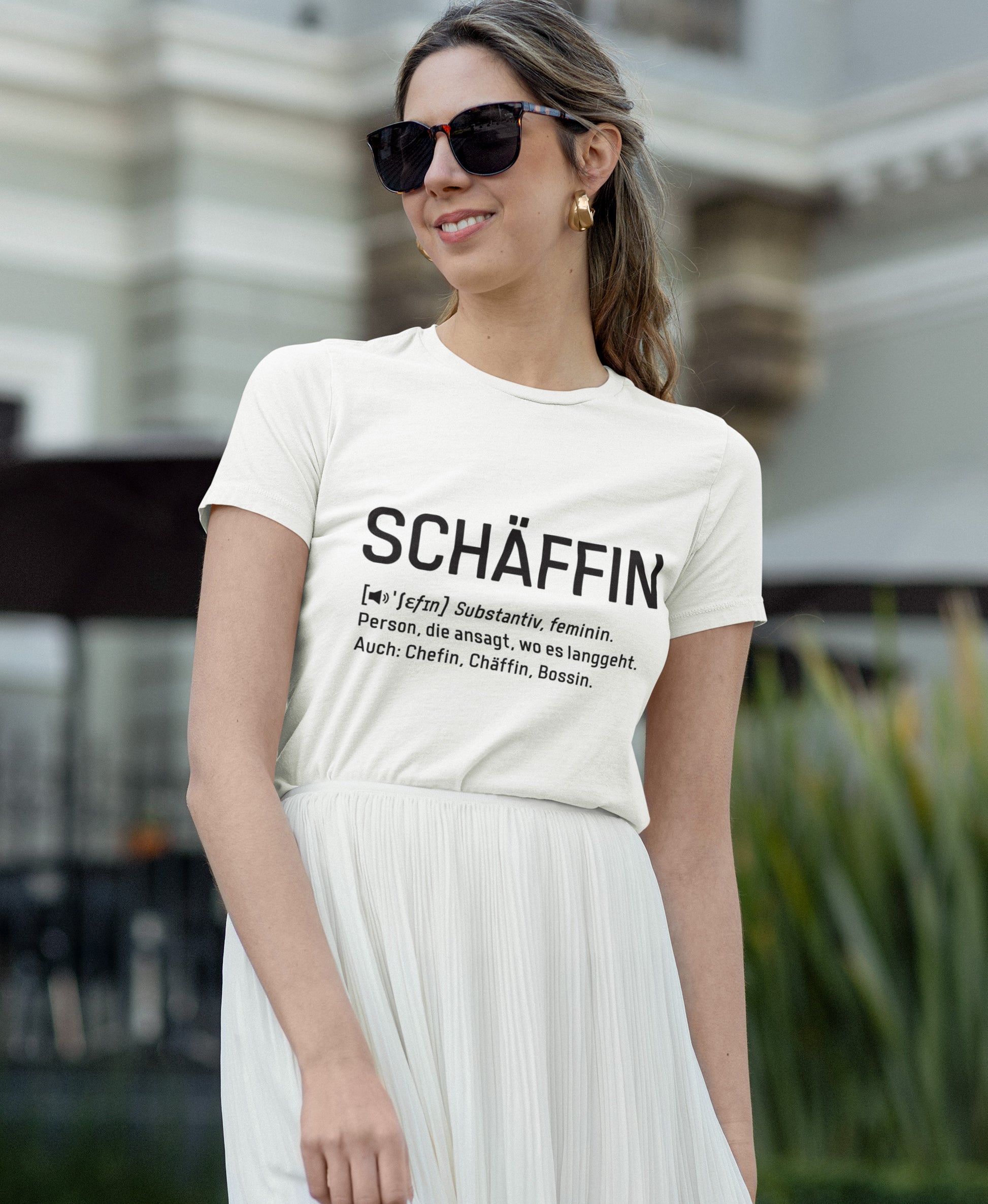 Junge Frau mit dunkler Sonnenbrille und weissem Rock trägt weisses SCHÄFFIN – Chefin Definition Bio T-Shirt mit schwarzn plakativen Frontaufdruck im Wörterbuch-Stil.