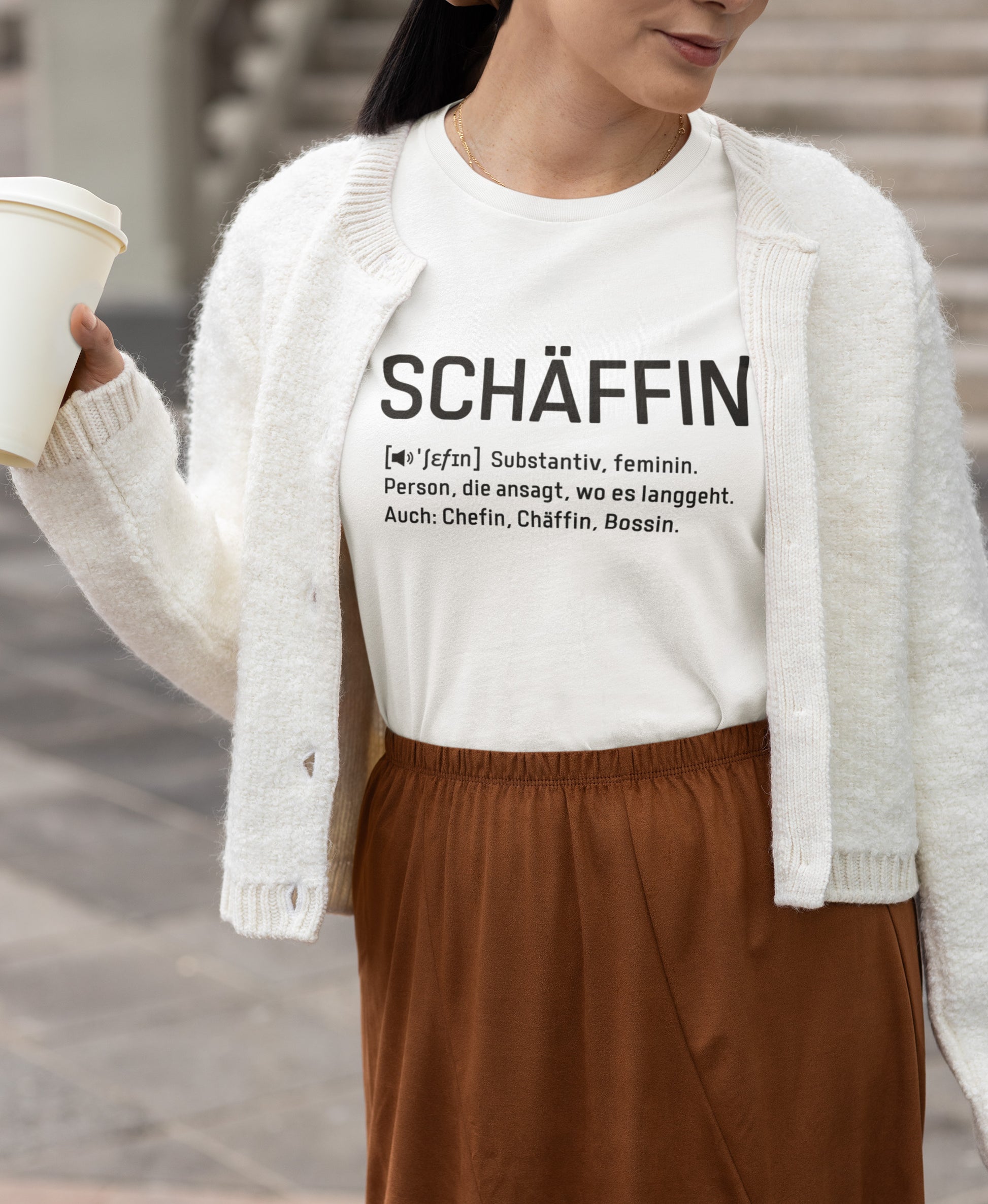 Frau mit Coffee-to-Go Becher trägt weissem Cardigan und schlichten Rock mit weissem SCHÄFFIN – Chefin Definition Bio T-Shirt mit schwarzen plakativen Frontaufdruck im Wörterbuch-Stil.
