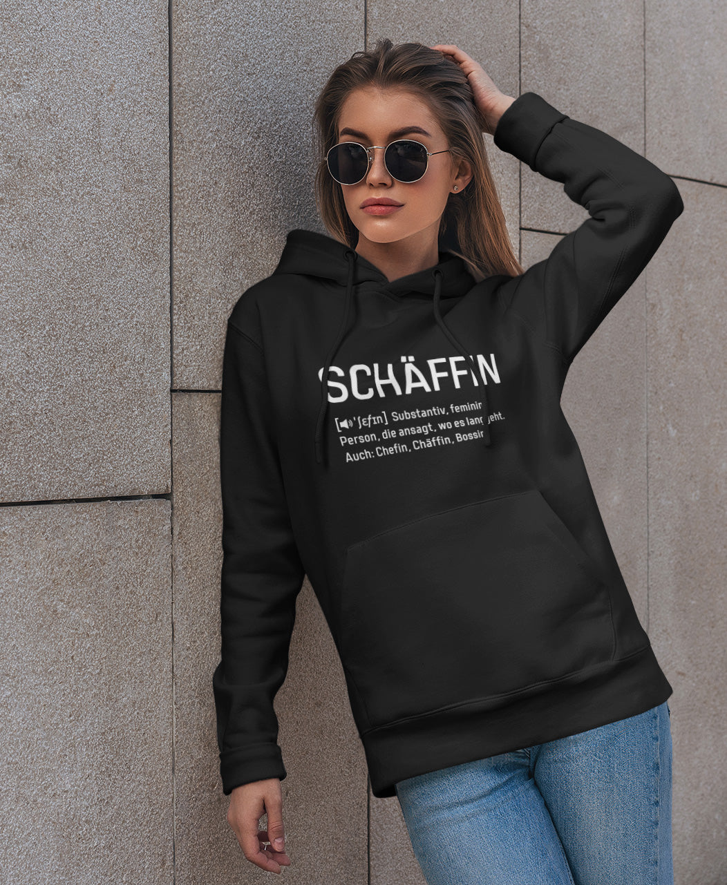 Frau mit Sonnenbrille lehnt an einer Hauswand und trägt SCHÄFFIN – Chefin Definition Bio-Hoodie in Farbe Schwarz