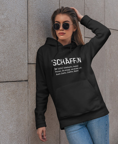 Frau mit Sonnenbrille lehnt an einer Hauswand und trägt SCHÄFFIN – Chefin Definition Bio-Hoodie in Farbe Schwarz